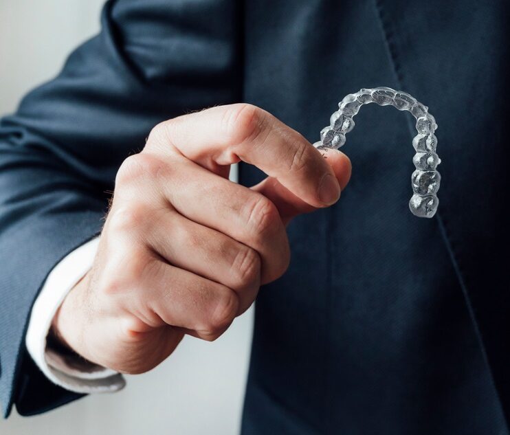 A person in a dark suit holding a transparent clear aligner tray between their fingers, showcasing an invisible orthodontic aligner used for teeth straightening.