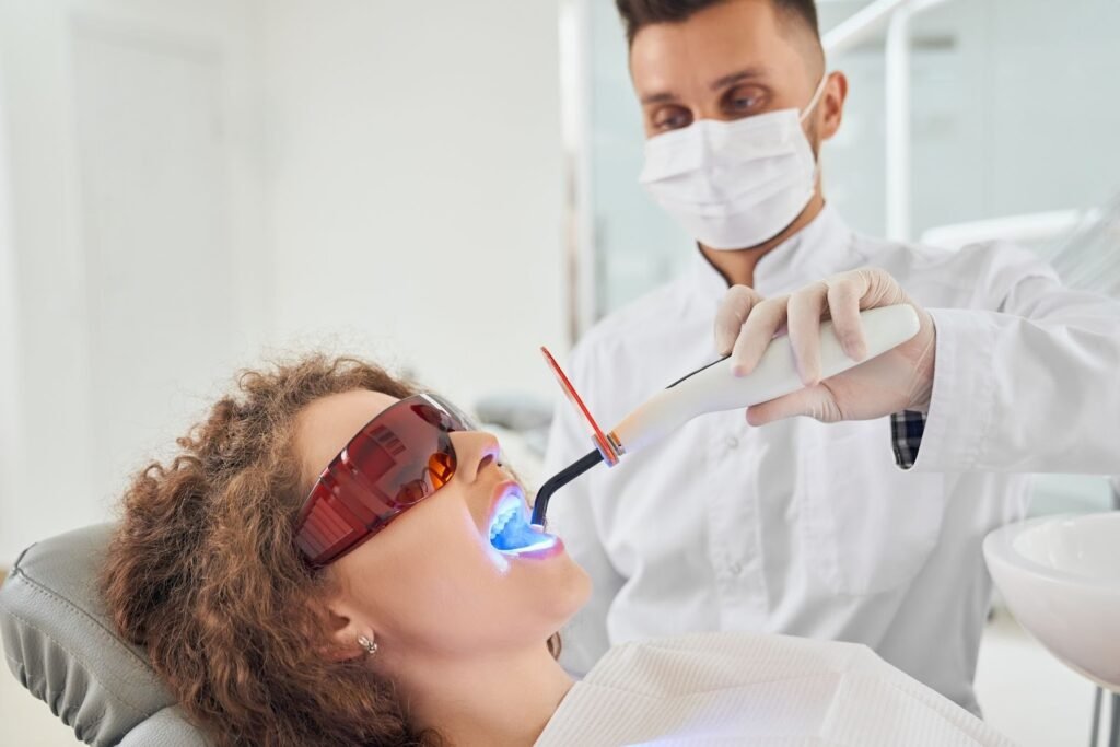 Dentist performing a teeth whitening procedure using a blue LED curing light on a patient wearing protective glasses in a dental clinic.