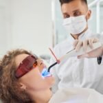 Dentist performing a teeth whitening procedure using a blue LED curing light on a patient wearing protective glasses in a dental clinic.