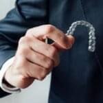 A person in a dark suit holding a transparent clear aligner tray between their fingers, showcasing an invisible orthodontic aligner used for teeth straightening.